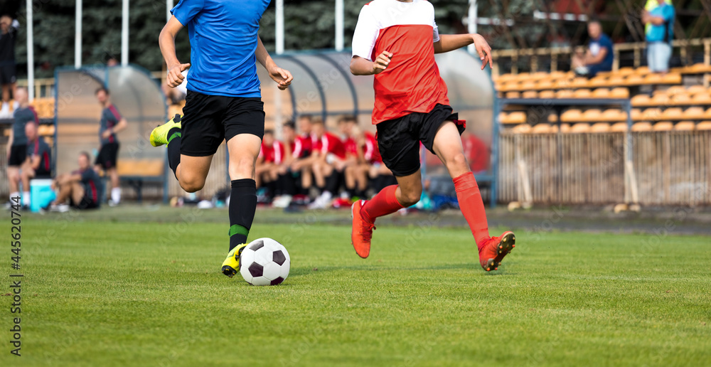 Two football players in running duel. Soccer teenage boys running fast with ball. Players sitting on substitute bench in the background. Footballers in red and blue jersey shirts Two football players in running duel. Soccer teenage boys running fast with ball. Players sitting on substitute bench in the background. Footballers in red and blue jersey shirts