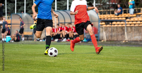 Two football players in running duel. Soccer teenage boys running fast with ball. Players sitting on substitute bench in the background. Footballers in red and blue jersey shirts