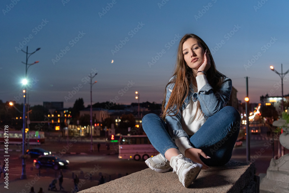 Fototapeta premium Beautiful young girl sits on evening city background. Stylish student girl lonely sitting on the high step