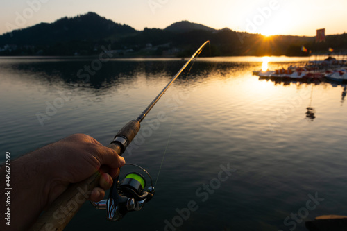 Hand holding fishing rod and a beautiful sunrise over calm lake with mountains in the distance. Colibita lake, Romania