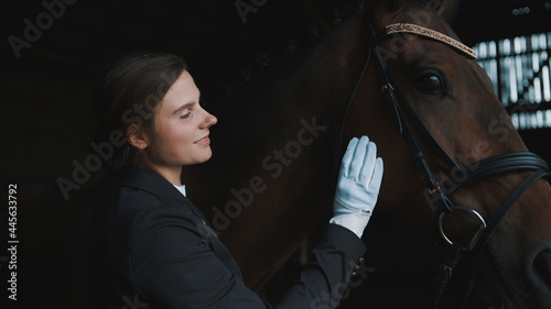 Female horse owner patting her seal brown horse in the stable. Sunlight striking through the window. The horse is wearing head jewelry. The girl is dressed in a black coat and wearing gloves.