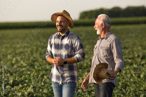 Two male farmers young and old standing in soy field talking laughing.