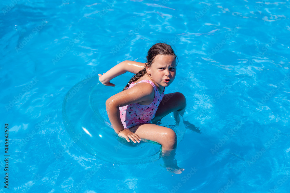 a five-year-old girl in a pink swimsuit is angry, that she can't keep her balance sitting on an inflatable circle in the hotel swimming pool. 