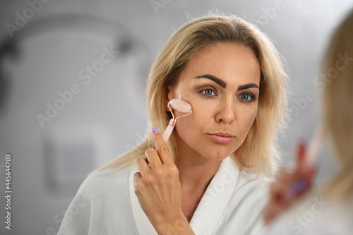 Gorgeous young woman looking at her mirror reflection and massaging her face with jade roller stone massager, performing lymphatic drainage facial massage