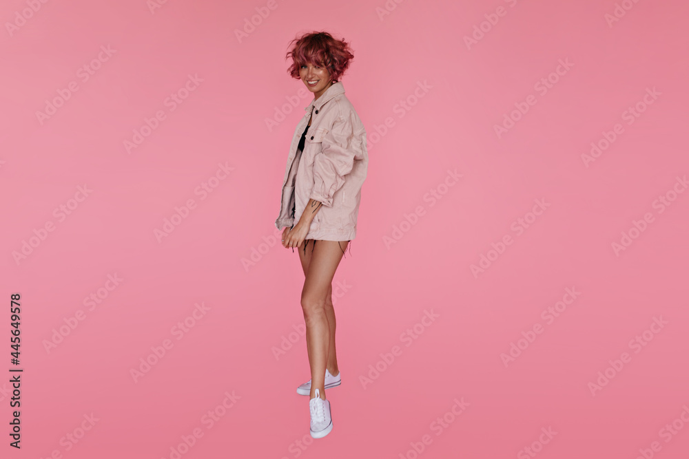 Charming happy girl in baggy denim beige jacket looks into camera. Full-length portrait of tanned curly woman smiling on isolated pink background. Charming happy girl in baggy denim beige jacket looks into camera. Full-length portrait of tanned curly woman smiling on isolated pink background.