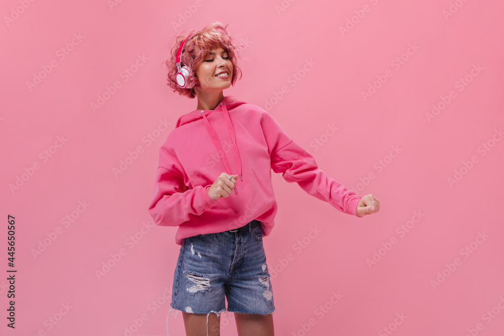 Pink-haired curly woman in headphones dances and listens to music. Portrait of pretty girl in pink hoodie and denim shorts on isolated . Pink-haired curly woman in headphones dances and listens to music. Portrait of pretty girl in pink hoodie and denim shorts on isolated .