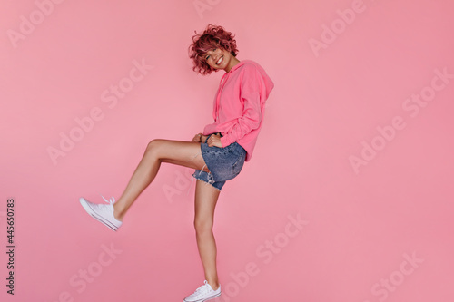 Happy woman moves on isolated. Charming curly girl in denim shorts and hoodie dances and smiles on pink background.