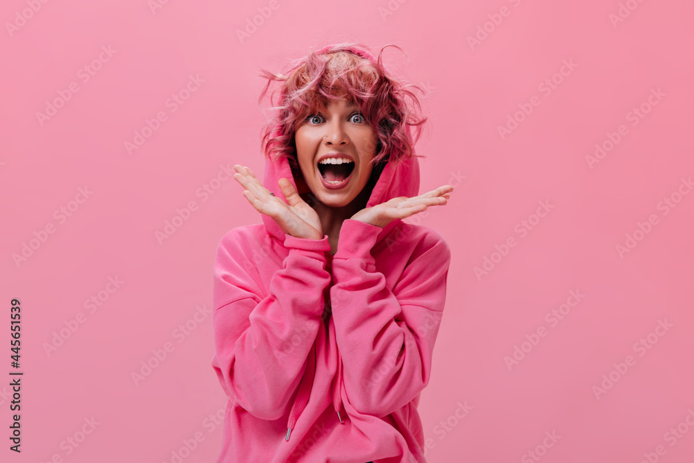 Shocked active woman in pink hoodie screams happily on isolated. Portrait of young curly girl looks surprised on pink background .