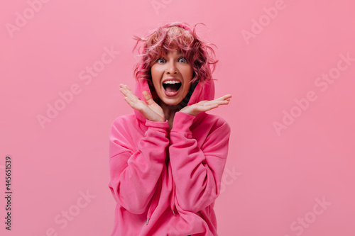 Shocked active woman in pink hoodie screams happily on isolated. Portrait of young curly girl looks surprised on pink background .
