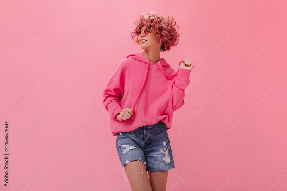 Curly woman in pink hoodie and denim shorts dance on isolated background. Happy cool girl in sunglasses moves and smiles on pink backdrop. Curly woman in pink hoodie and denim shorts dance on isolated background. Happy cool girl in sunglasses moves and smiles on pink backdrop.