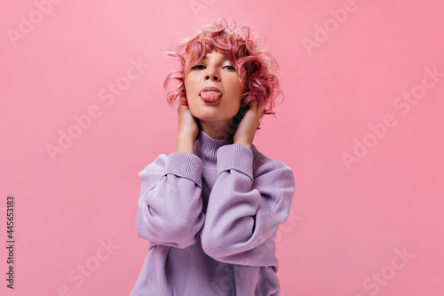 Happy young curly pink-haired woman demonstrates tongue. Pretty girl in purple sweater makes funny face on isolated.