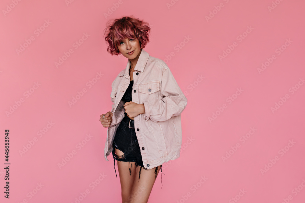 Attractive curly woman in denim pink jacket smiling on isolated. Short-haired tanned girl in black shorts and cropped tee poses on pink background. Attractive curly woman in denim pink jacket smiling on isolated. Short-haired tanned girl in black shorts and cropped tee poses on pink background.