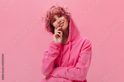 Close-up portrait of curly pink-haired girl in pink hoodie looking into camera with sincere smile. Pretty woman poses on isolated background. © Look!
