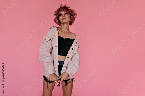 Curly pink-haired woman in dark denim shorts and stylish oversized jacket poses on pink background. Attractive tattooed girl in sunglasses looks gorgeous .