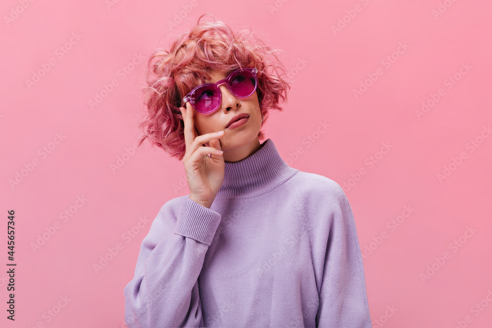 Portrait of pink-haired curly thoughtful woman in purple sweater and fuchsia sunglasses posing on isolated background .