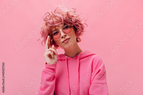 Attractive woman in stylish sunglasses looks into camera. Pretty curly girl in bright fuchsia color hoodie poses on isolated pink background.