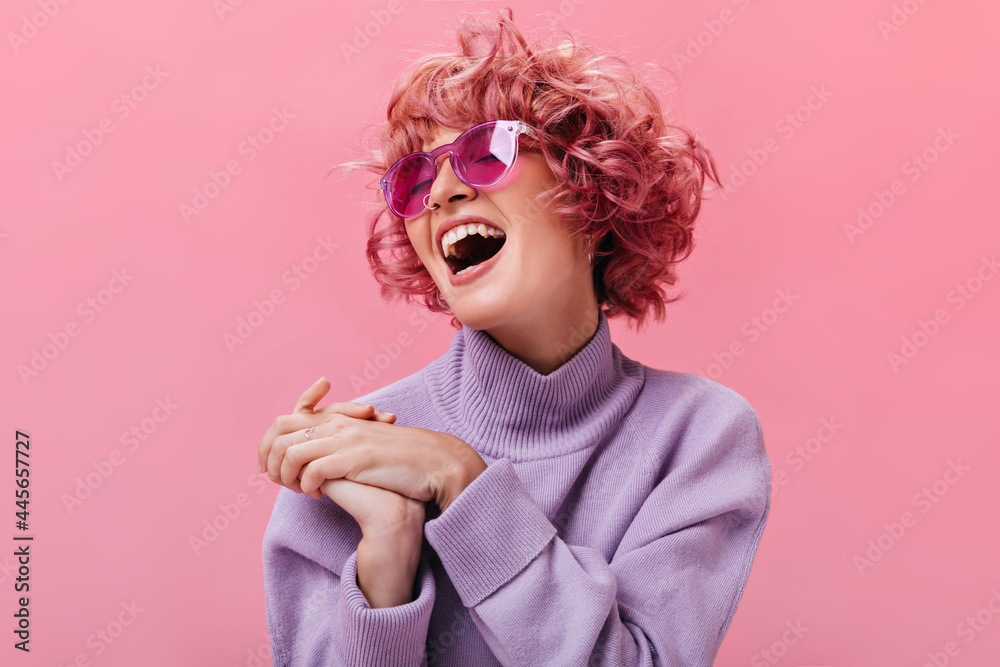 Young good-humored curly woman in fuchsia sunglasses and purple cashmere sweater laughs on isolated pink background. Young good-humored curly woman in fuchsia sunglasses and purple cashmere sweater laughs on isolated pink background.