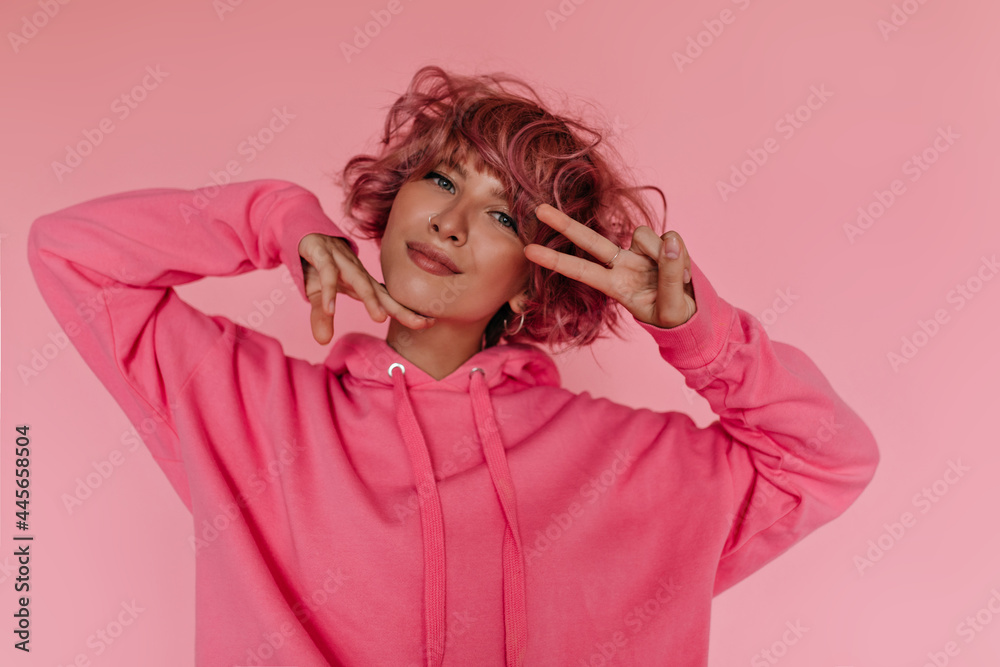 Tanned curly pink-haired woman shows peace sign on isolated. Portrait of happy girl in bright hoodie poses on pink background. Tanned curly pink-haired woman shows peace sign on isolated. Portrait of happy girl in bright hoodie poses on pink background.