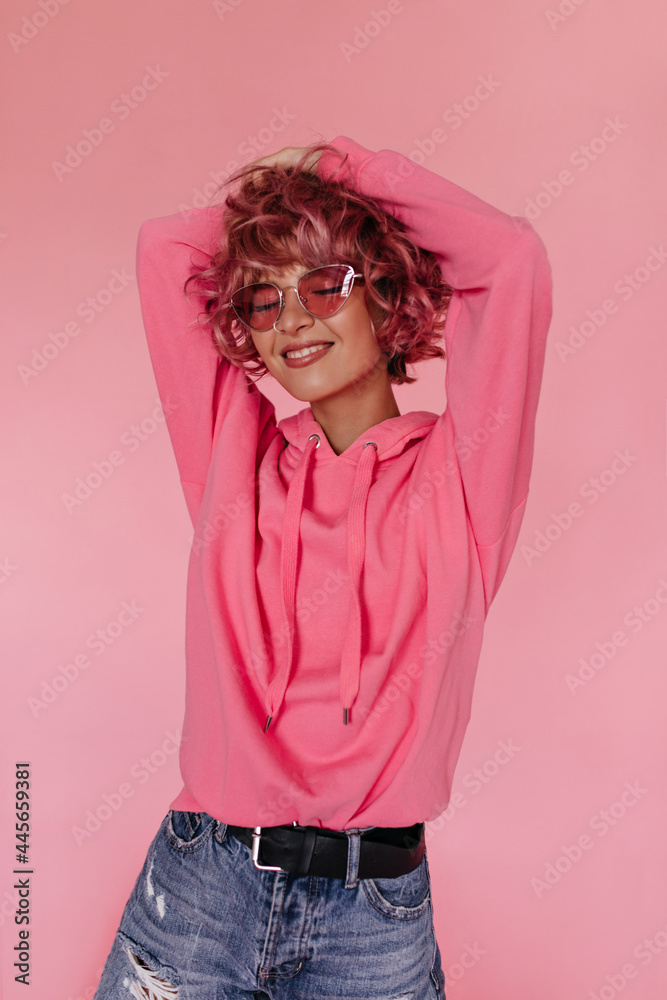 Charming curly pink-haired girl in bright sunglasses and fuchsia hoodie smiles widely and poses on isolated background in good mood. Charming curly pink-haired girl in bright sunglasses and fuchsia hoodie smiles widely and poses on isolated background in good mood.