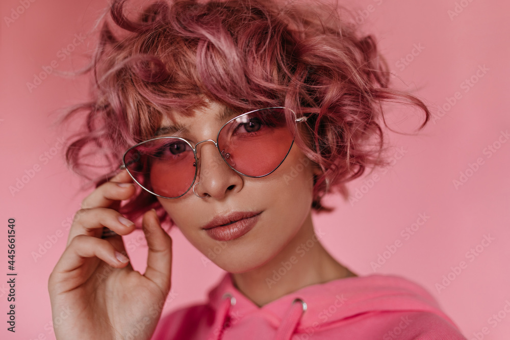 Close-up portrait of curly woman in sunglasses looking into camera. Charming girl in pink hoodie poses on isolated background.