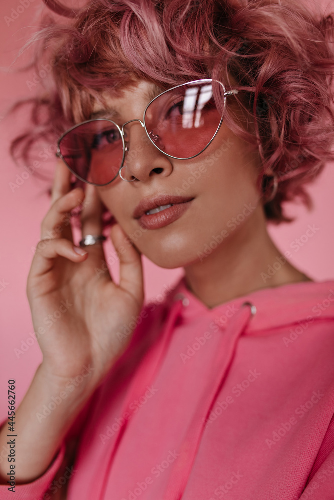 Attractive short-haired curly woman in pink sunglasses looks into camera. Close-up inside portrait of pretty girl in hoodie posing on isolated.