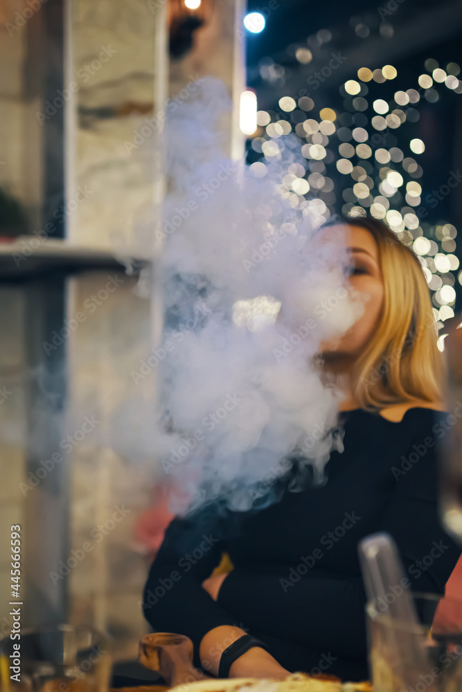Woman smokes hookah in lounge bar. Female in black dress is resting at cafe table. Bright lights on background. Thick smoke from shisha.