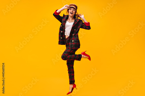 Beautiful lady in stylish suit, cap and eyeglasses jumping on orange background. Happy woman in modern outfit and red shoes posing..