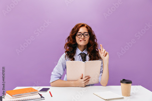 Red haired girl crossed fingers and holds notebook.Curly young woman in round eyeglasses and stylish school outfit posing with book..