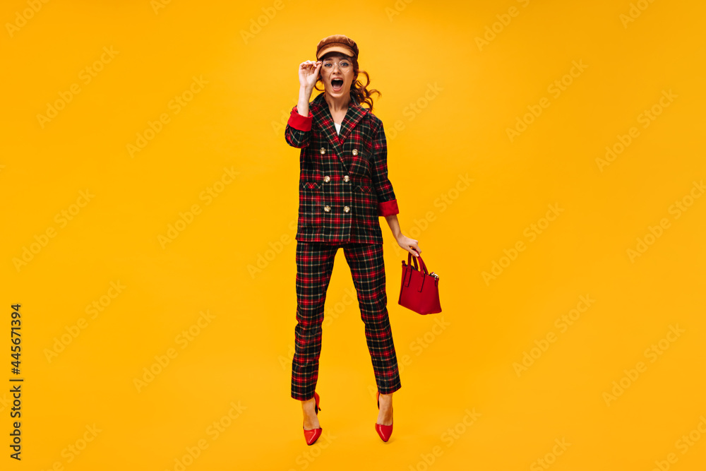 Full length woman in plaid outfit jumps on orange background and holds handbag. Charming girl in eyeglasses and brown cap posing.. Full length woman in plaid outfit jumps on orange background and holds handbag. Charming girl in eyeglasses and brown cap posing..