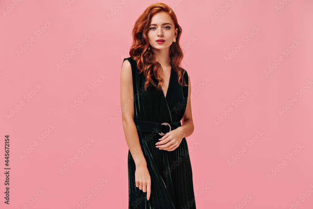Ginger woman in green dress poses on pink background. Red-haired curly lady in stylish velvet outfit looking into camera.. Ginger woman in green dress poses on pink background. Red-haired curly lady in stylish velvet outfit looking into camera..
