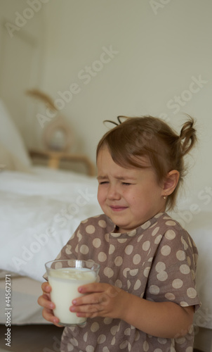 a cute little girl is holding a glass glass of kefir milk in her hands and is wrinkled does not want to drink disgusting