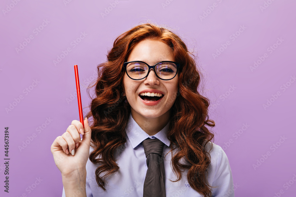 Portrait of woman in shirt smiling and holding red pencil. Joyful girl with ginger curly hair in modern outfit laughs on isolated background..