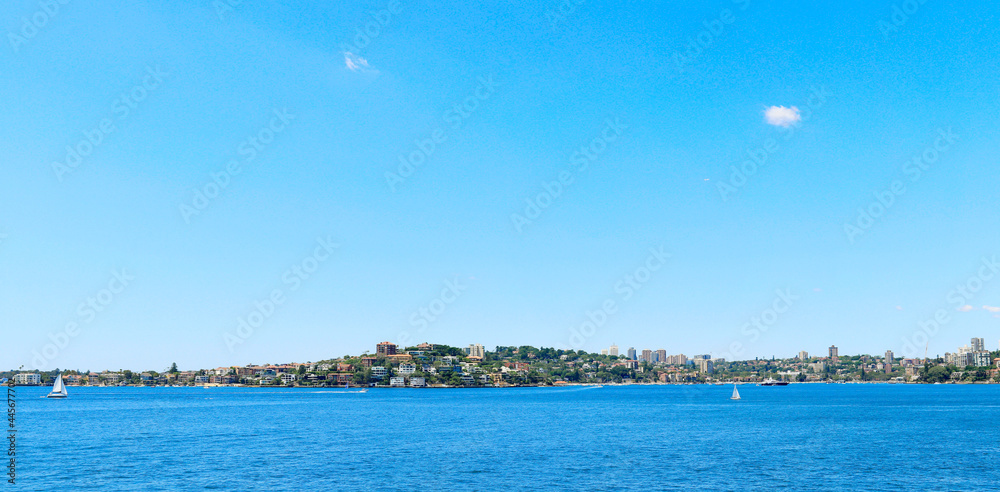 Under blue sky and white clouds, sailing boats driving on blue sea; a city by the sea in the distance. Panoramic shot
