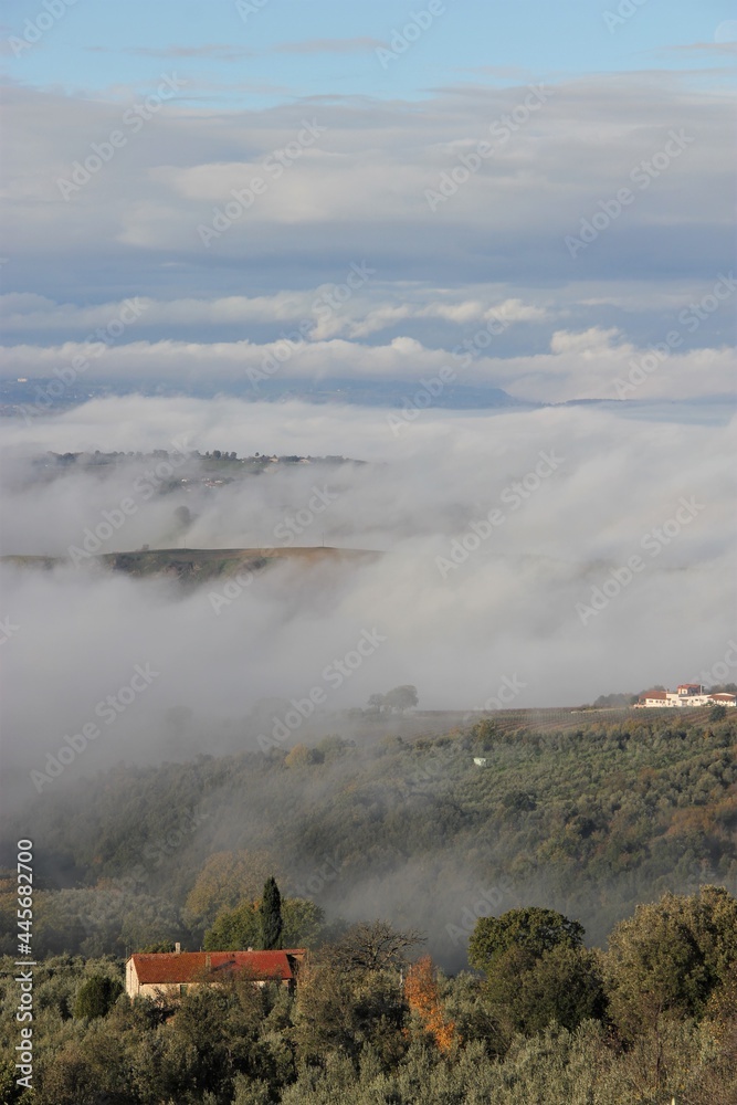 The green rolling hills of Umbria, Italy, a quaint maroon roofed country house in the foreground, submerged in towering thick grey, blue and white clouds, on a late autumn morning