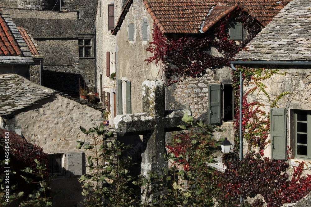 The grey, petrified world of the Medieval Southern French hamlet of La Couvertoirade, dominated by a mighty stone cross, a maze of stone buildings overgrown with red vine leaves in the back