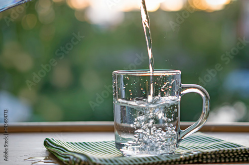 Drinking water is poured into a glass with a handle on a wooden table from a glass jug against the background of green nature.Sunset. The topic is thirst quenching, ecology. Selective focus