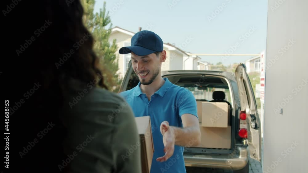 Friendly delivery worker is bringing carton box to female client signing papers talking standing at front door in suburban area. People and commerce concept.