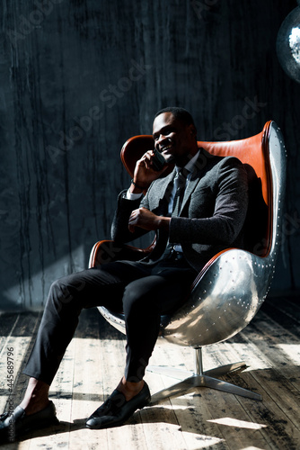 Handsome african american businessman in a jacket and trousers sits on a chair and looks away smiling