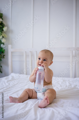 a baby girl in a diaper holds a bottle of water and sits on a white bed