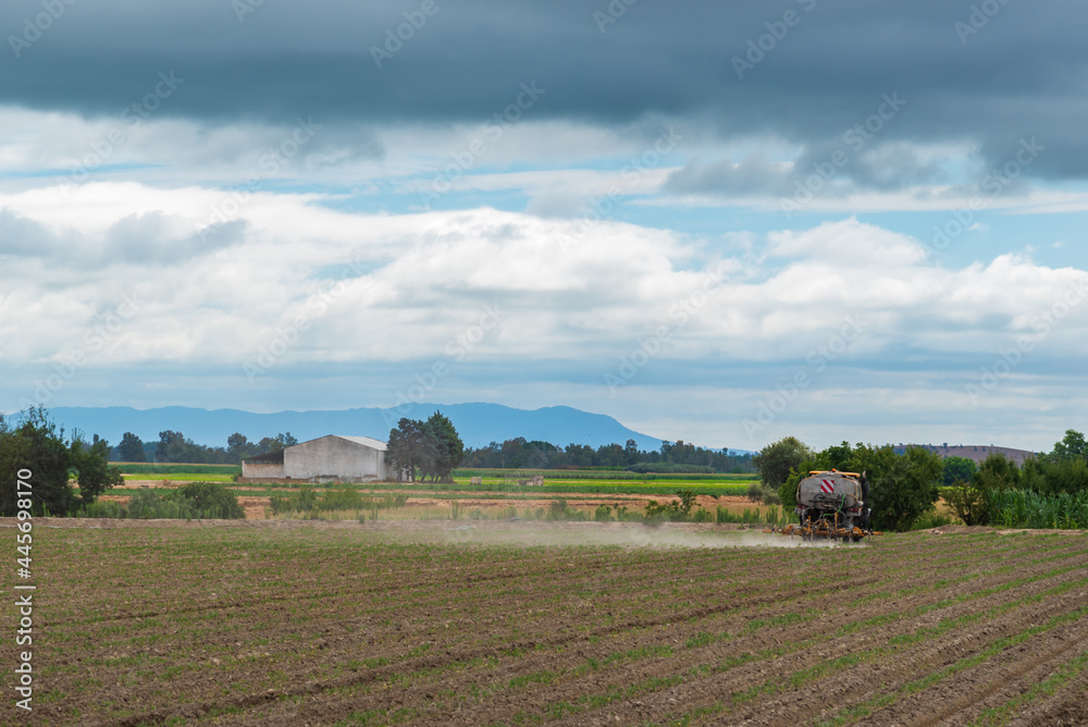 Agricultural tractor working in the field, with a green landscape, an agricultural industrial warehouse, mountains and the cloudy sky. Agricultural tractor working in the field, with a green landscape, an agricultural industrial warehouse, mountains and the cloudy sky.