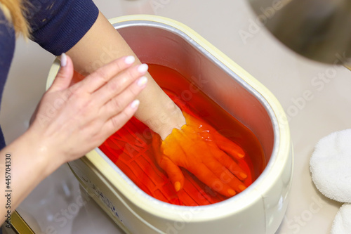 Paraffin theoraphy. Female hand and orange paraffin wax in bowl. Manicure and skincare. Woman girl in beauty spa salon.
