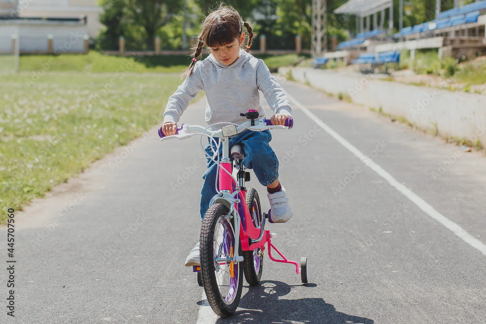 little cute girl 4 years old with pigtails learning to ride a bike in a sports stadium in summer little cute girl 4 years old with pigtails learning to ride a bike in a sports stadium in summer