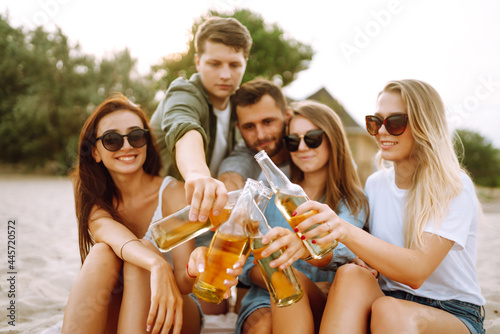 Group of friends cheers and drink beers on the beach. Young friends sitting together sitting near the sea and enjoy summer party. Beach holiday and summer vacation concept.