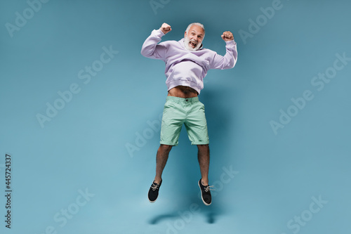 Happy man in hoodie jumping on blue background. Cheerful guy with white beard with tattoos in fashionable clothes posing..