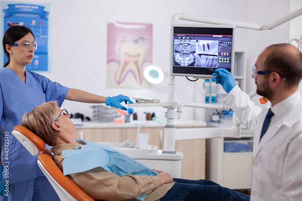 Fototapeta premium Stomatolog explaining diagnosis to senior woman during dental examination. Medical teeth care taker pointing at patient radiography on screen sitting on chair.