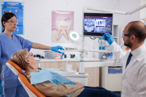 Stomatolog explaining diagnosis to senior woman during dental examination. Medical teeth care taker pointing at patient radiography on screen sitting on chair.