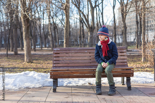 Caucasian boy sits in a winter park on a bench alone. The boy is sad. Snow on the ground in the background