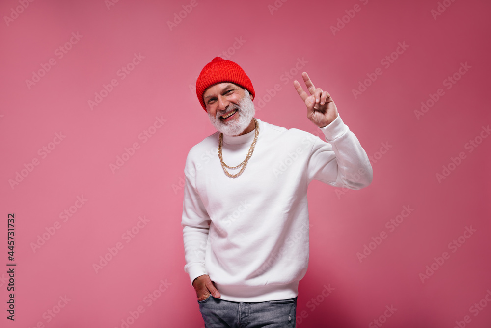 Happy man in orange hat and white sweater showing peace sign on pink background. Adult handsome guy in modern clothes smiling..