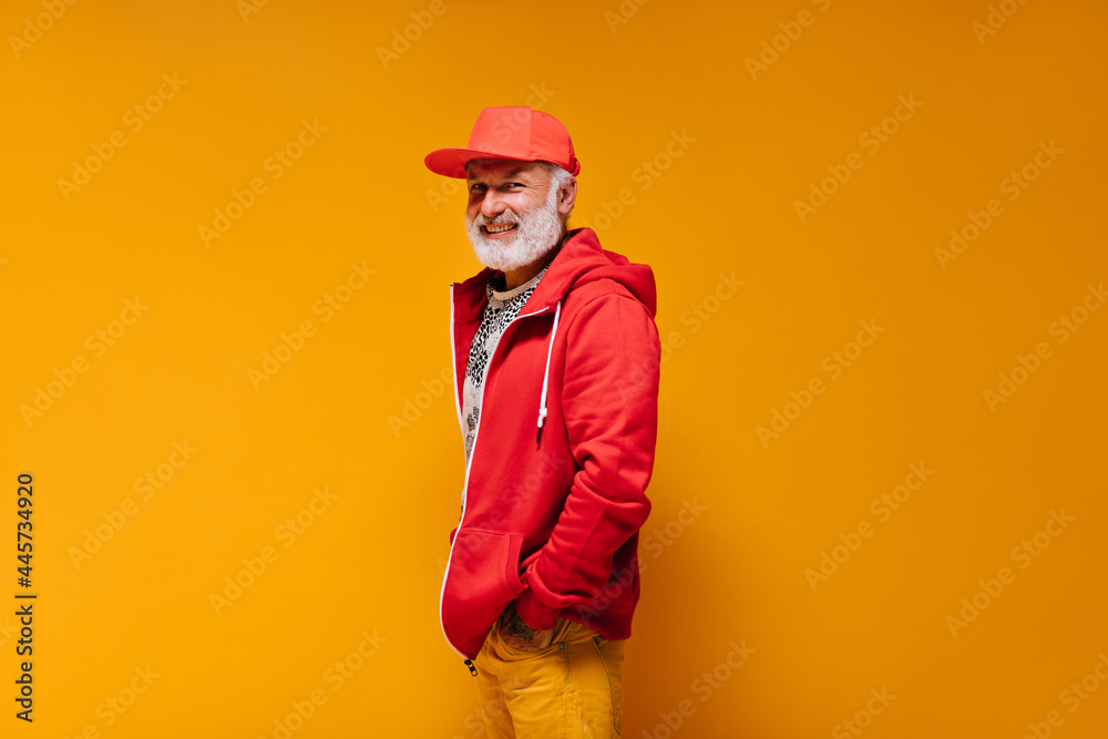Smiling man in red outfit looks into camera on orange background. Trendy guy with grey beard in bright cap and sweatshirt looking into camera..