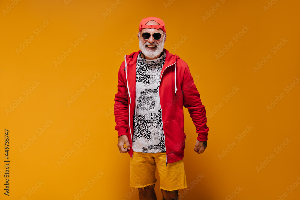 Charming man in red outfit smiling and posing on orange background. Cheerful adult guy with white hair in cap and sunglasses laughs.. Charming man in red outfit smiling and posing on orange background. Cheerful adult guy with white hair in cap and sunglasses laughs..